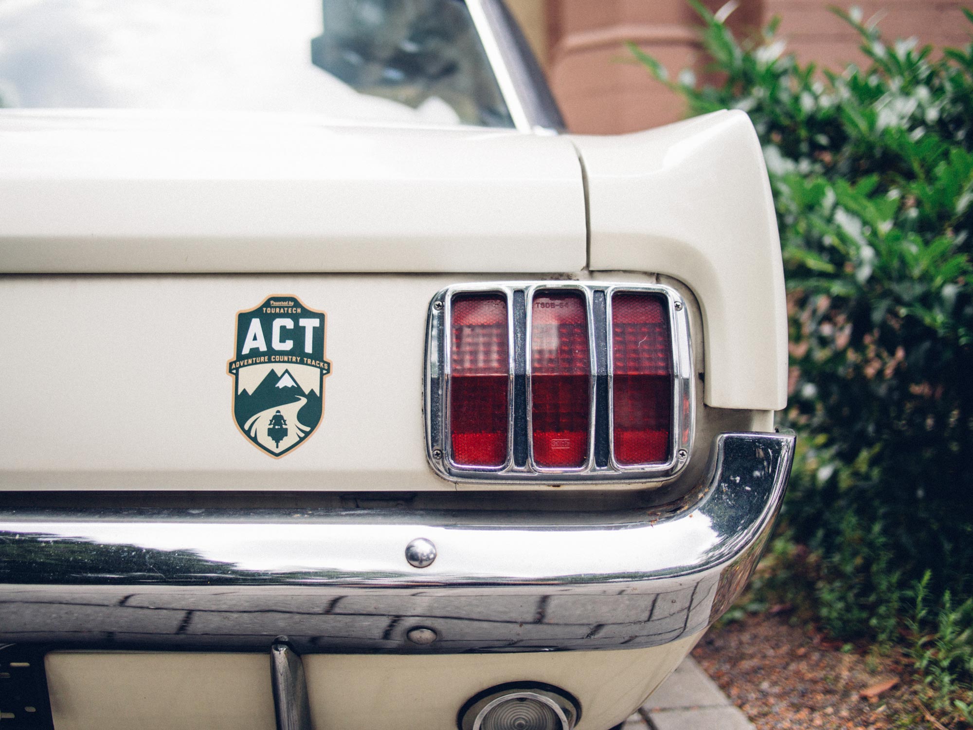 Man sieht das Foto eines Ford Mustang aus den 60er Jahren. Ein Aufkleber mit dem Logo der Adventure Country Tracks von TOURATECH klebt neben dem prägnanten Rücklicht. Das Logo hat eine Wappenform, oben sind die Buchstaben 