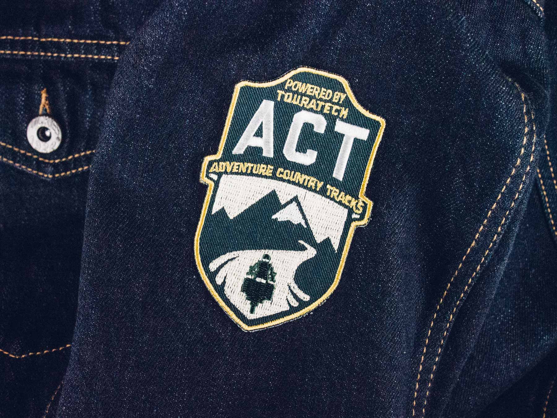 Man sieht das Foto einer Jeansjacke mit Logo-Aufnäher der Adventure Country Tracks von TOURATECH. Das Logo hat eine Wappenform, oben sind die Buchstaben 