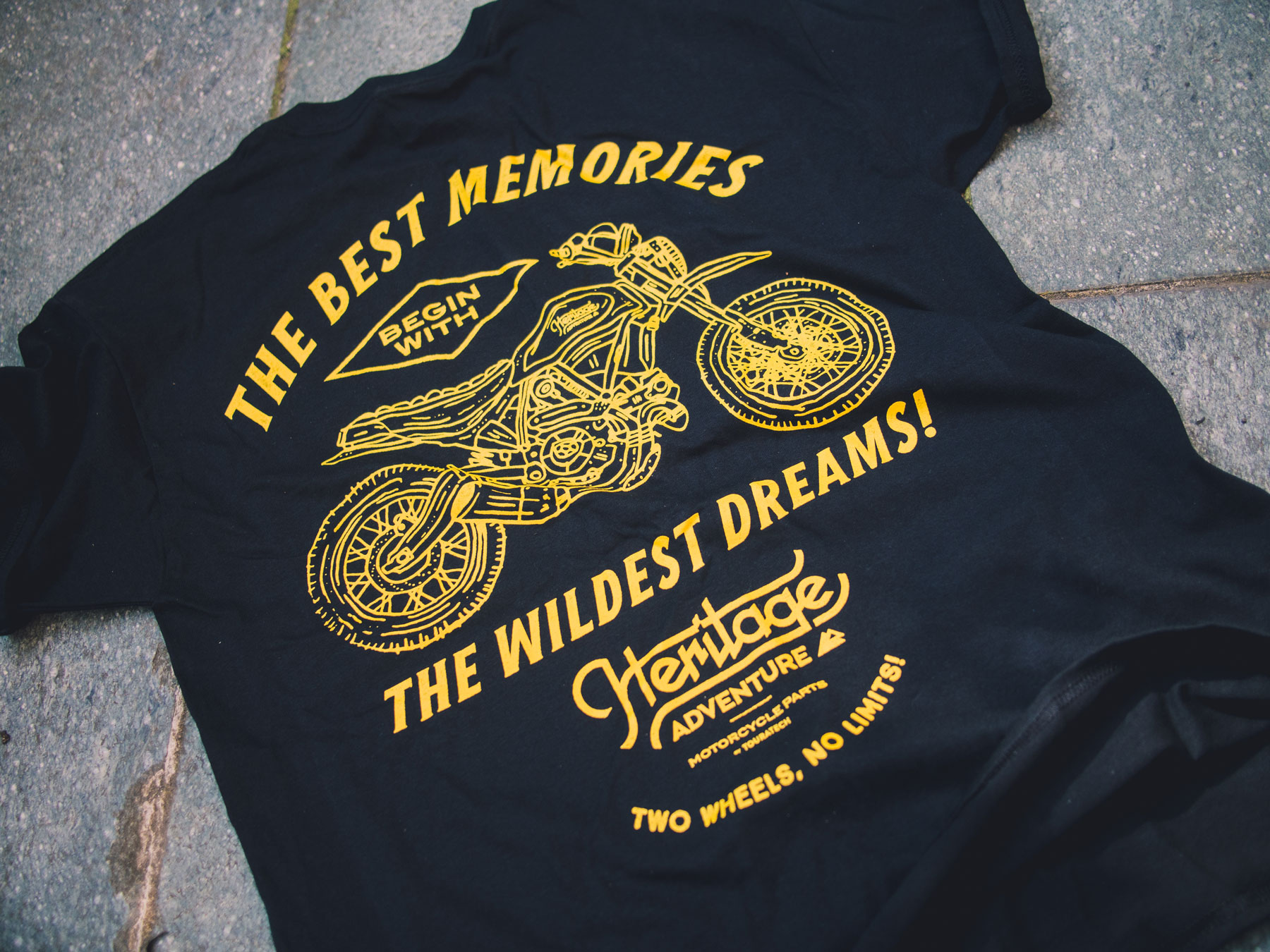 Man sieht ein T-Shirt von Heritage Adventure von TOURATECH. Auf der Rückseite ist eine gezeichnete Ducati Scrambler sowie der Spruch 
